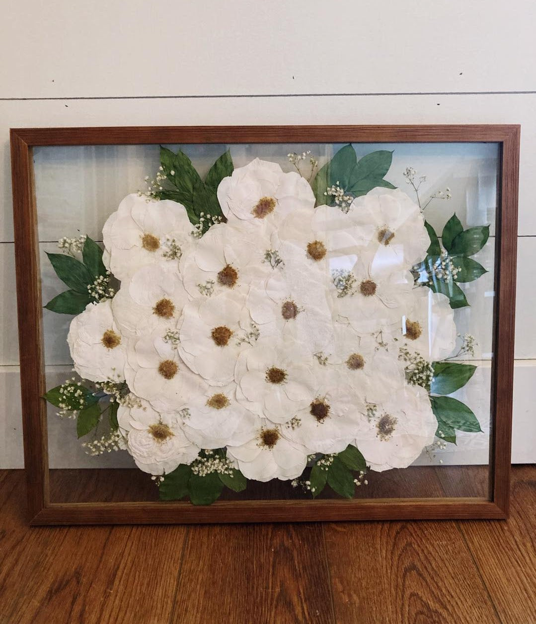 Framed arrangement of white flowers with green leaves on a wooden floor.